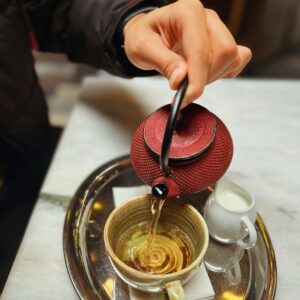 A hand pours tea from a red teapot into a cup on a tray, in an İzmir cafe.