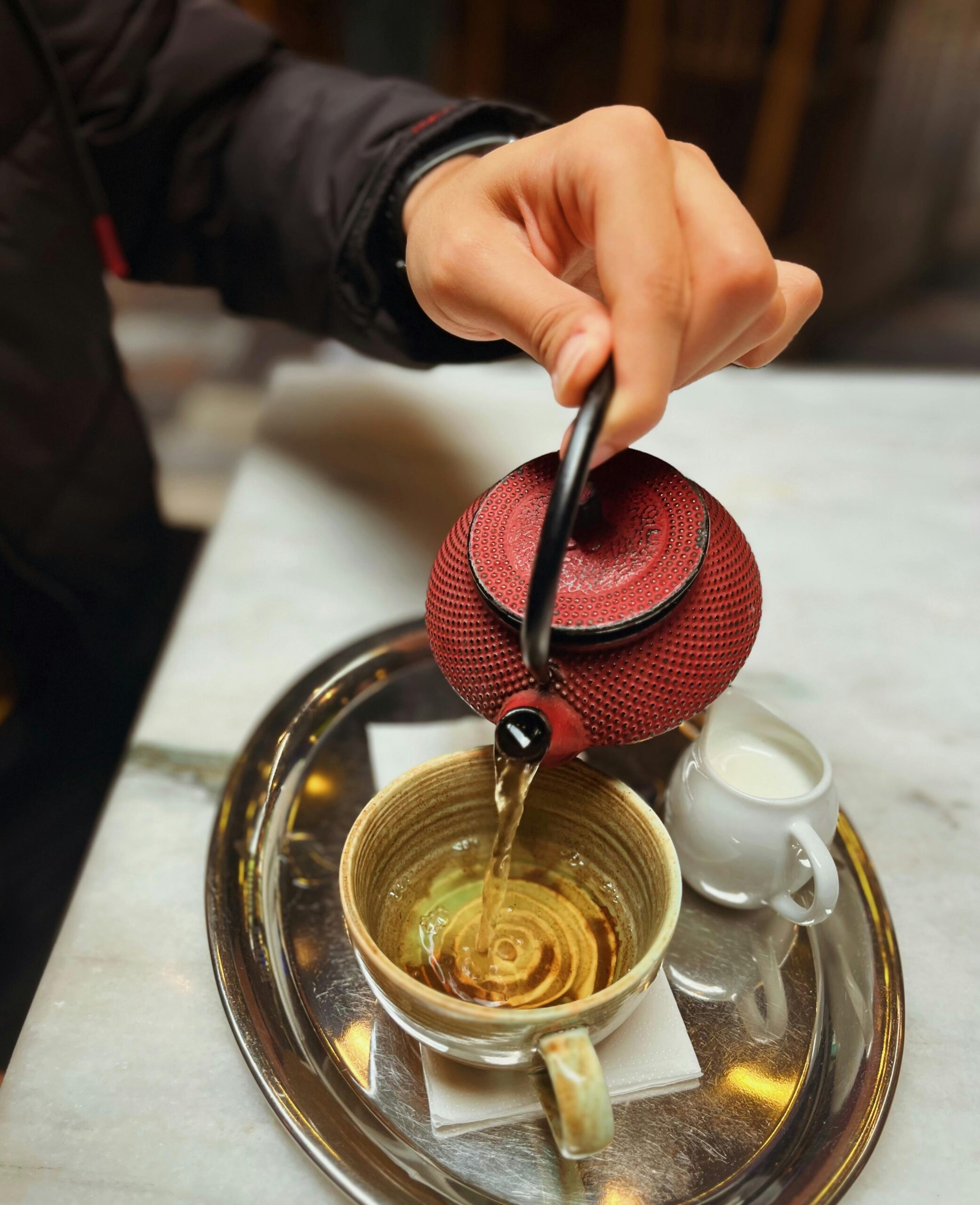 A hand pours tea from a red teapot into a cup on a tray, in an İzmir cafe.